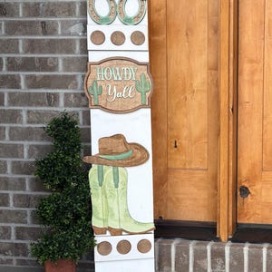 May include: A white wooden porch sign with a western theme. It features a horseshoe design, the words "Howdy Y'all", a cowboy hat, and cowboy boots. The sign is decorated with brown, green, and white accents.