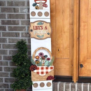 May include: A white wooden sign with a mushroom and ladybug design at the top. The sign reads "Life's a Picnic" and features a basket of flowers and ladybugs. The sign is decorated with a red and white checkered pattern.