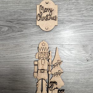 May include: Wooden cutout of a nutcracker holding a staff, standing next to a Christmas tree and a gift. The cutout is on a woodgrain background. A separate wooden cutout with the text "Merry Christmas" is above the nutcracker.