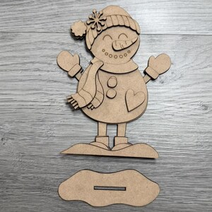 May include: A wooden snowman figure with a hat, scarf, and mittens. The snowman has a carrot nose, button eyes, and a heart on its belly. It stands on a base with a slot for display. A festive winter decoration.