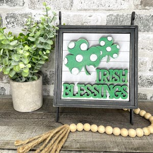 May include: A decorative sign with a black frame features green shamrocks and the words "Irish Blessings." The sign is displayed on a stand, with a string of wooden beads and a small potted plant in the background. The sign is a St. Patrick's Day decoration.