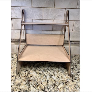 May include: A two-tiered wooden shelf unit with a triangular frame. The shelf is made of light brown wood with a dark brown border.