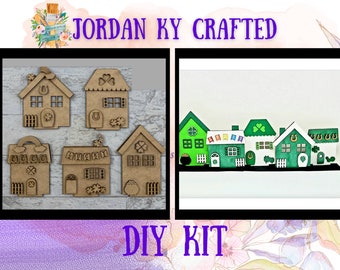 May include: A DIY kit for crafting a miniature Irish village. The kit includes five wooden houses with shamrock and horseshoe details. The houses are shown assembled in the second image, painted green and white with rainbow flags and gold coins.