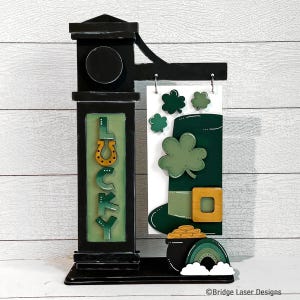 May include: A wooden sign with a black frame and a green and gold design. The sign says "LUCKY" and features a leprechaun's hat, a pot of gold, and a rainbow.