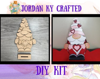 May include: A Valentine's Day gnome DIY kit. The kit includes a wooden gnome with a red hat, white beard, and pink hearts. The gnome is holding a large pink heart in its hands. The kit also includes a wooden base for the gnome to stand on. The gnome is shown in both its unfinished and finished states.