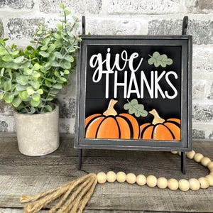 May include: A black framed sign with the words "give THANKS" in white script, featuring orange pumpkins and green leaves. A small potted plant and a wooden bead garland are also in the image.