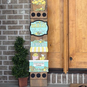 May include: Wooden lemonade stand sign with a basket of lemons, the phrase "Squeeze the Day", and the word "Lemonade". The sign also features a pink bucket of lemons, a pitcher of lemonade, and the text "Farm Fresh Lemons".