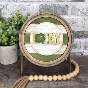 May include: A round wooden sign with the word "LUCKY" in gold lettering and a green shamrock. The sign has a white and green background and is surrounded by a wooden frame. A wooden stand and a string of wooden beads are also visible.
