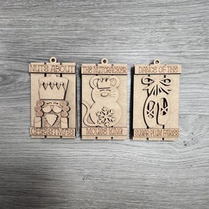May include: Three wooden ornaments with a Nutcracker theme. The first ornament says "Nuts About Christmas" and features a Nutcracker soldier. The second ornament says "The Nutcracker Mouse King" and features a mouse wearing a crown. The third ornament says "Dance of the Sugar Plum Fairies" and features a pair of ballet slippers.