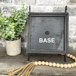 May include: A black wooden sign with the word "BASE" in white lettering. The sign is framed in a dark metal and has a small hole in the center. A small potted plant and a wooden bead garland are also in the image.