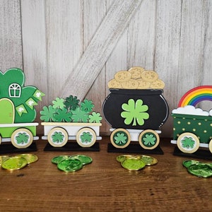 May include: A set of four wooden train cars decorated for St. Patrick's Day. The cars feature shamrocks, a pot of gold, a rainbow, and a house. Each car has wheels and is painted in green, gold, and black.