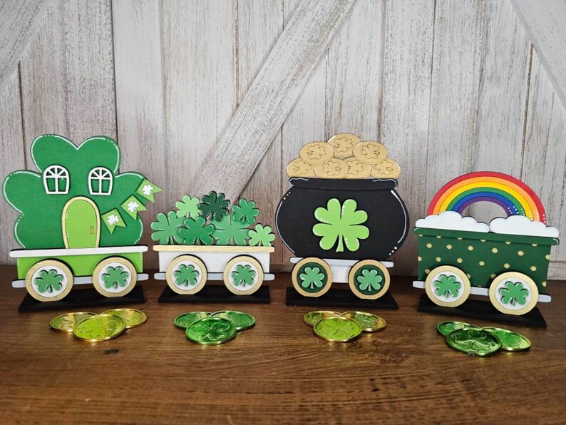 May include: A set of four wooden train cars decorated for St. Patrick's Day. The cars feature shamrocks, a pot of gold, a rainbow, and a house. Each car has wheels and is painted in green, gold, and black.