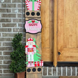 May include: A wooden Christmas sign with a pink and green nutcracker, a pink gift box with candy canes, and a pink sign that says "Happy Holidays".