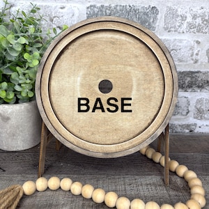 May include: A round, wooden base with the word "BASE" in black block letters. The base is supported by small wooden legs. A strand of wooden beads and a small plant in a pot are in the foreground.