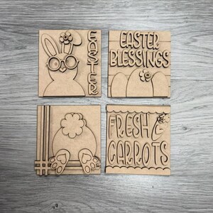 May include: Four square wooden Easter decorations. One features a bunny with glasses and the word "Easter". Another says "Easter Blessings" with floral accents. The third shows a bunny's backside, and the last says "Fresh Carrots".