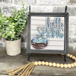 May include: A decorative wooden sign with the words "hello SPRING" in blue script. The sign features a nest with three speckled eggs. A small potted plant and a wooden bead garland are also in the image.