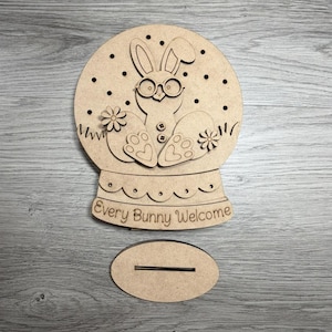 May include: A wooden Easter-themed snow globe with a bunny wearing glasses, flowers, and the text "Every Bunny Welcome." Includes a separate oval base. The design is laser-cut from light brown wood.