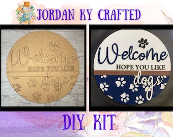 May include: DIY craft kit for a welcome sign with the text "Welcome Hope You Like Dogs" in a circle with paw prints. The sign is shown in two versions, one with a natural wood finish and the other with a blue and white paw print design.