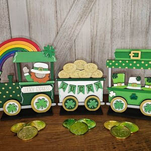 May include: A wooden train set with three carriages decorated for St. Patrick's Day. The first carriage is a green engine with gold polka dots and a rainbow on top. The second carriage is white with the word "LUCKY" in green letters and a pile of gold coins. The third carriage is green with gold polka dots and has three leprechaun hats on top. The train is on a wooden table with gold coins scattered around.