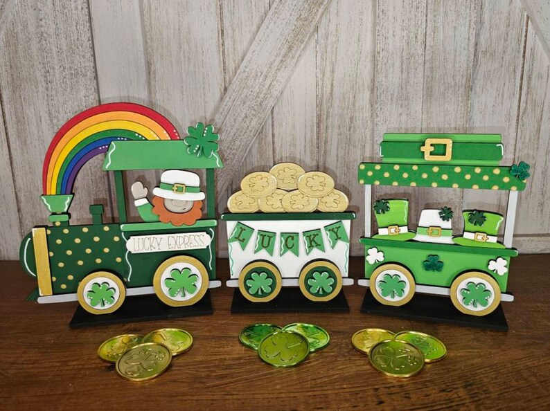 May include: A wooden train set with three carriages decorated for St. Patrick's Day. The first carriage is a green engine with gold polka dots and a rainbow on top. The second carriage is white with the word "LUCKY" in green letters and a pile of gold coins. The third carriage is green with gold polka dots and has three leprechaun hats on top. The train is on a wooden table with gold coins scattered around.