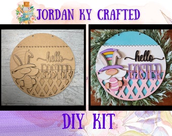 May include: DIY kit for a wooden Easter wreath featuring a gnome holding an Easter egg. The wreath is decorated with a pink and blue background, a white banner with the text "hello easter", and a diamond-shaped lattice pattern.