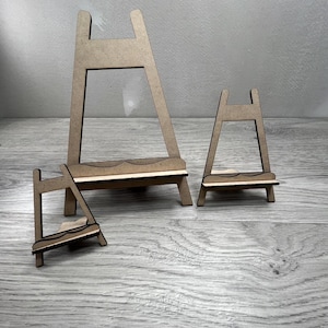 May include: Three small, unfinished wooden easels of varying sizes. The easels are made of light brown wood and have a simple A-frame design with a small shelf. They are arranged on a light gray wooden surface.
