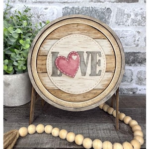 May include: A round wooden sign with the word "LOVE" and a pink heart. The sign has a rustic, farmhouse style with a wooden bead garland and a small plant in the background. The sign is supported by a wooden stand.