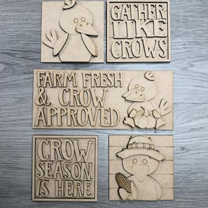 May include: A set of five unfinished light brown wooden signs featuring laser-cut designs. The signs include illustrations of crows, a pumpkin, and corn, along with the text GATHER LIKE CROWS, FARM FRESH & CROW APPROVED, and CROW SEASON IS HERE.