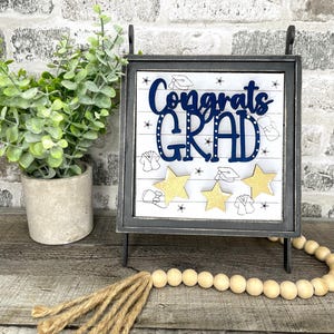 May include: A graduation-themed sign with the words "Congrats GRAD" in navy blue, accented with gold stars and graduation cap illustrations. The sign is set in a black metal frame and stands on a wooden surface.