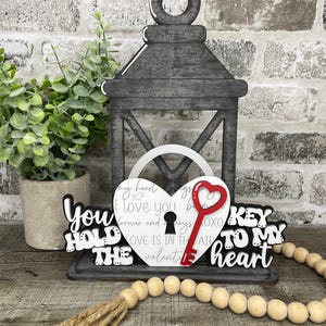 May include: A decorative wooden lantern with a heart-shaped cutout and a red key. The words "You Hold The Key To My Heart" are displayed. A wooden bead garland and a potted plant are also present.