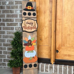 May include: A wooden Thanksgiving sign with a turkey, pumpkins, and a pilgrim hat. The sign says "Happy Thanksgiving".