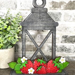 May include: A gray wooden lantern with a decorative strawberry base. The strawberries are red with white speckles, green leaves, and white flowers. The lantern has an open design and a ring at the top.