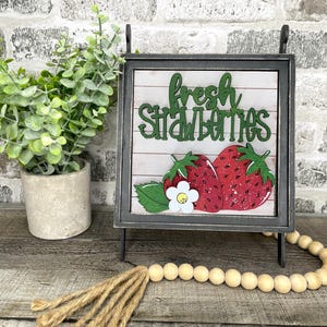 May include: A decorative sign with the words "fresh strawberries" in green script. The sign features two red strawberries with green leaves and a white flower. The sign is in a black frame and is displayed on a wooden surface.