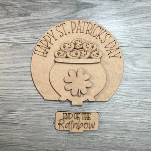 May include: A wooden cutout of a pot of gold at the end of the rainbow. The pot is filled with gold coins and has a shamrock on the side. The text "Happy St. Patrick's Day" is written around the top of the pot. The text "End of the Rainbow" is written on a separate wooden cutout.