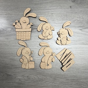 May include: A collection of wooden Easter bunny cutouts in various poses. One bunny holds carrots, another an egg, and one is in a basket. The cutouts are light brown and feature simple, cartoon-like designs, perfect for Easter crafts.