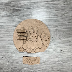 May include: A wooden cutout of a bunny driving a carrot car with a signpost that reads "Egg Hunt", "Carrots", and "Bunny". The signpost is on a round wooden base with clouds and the words "Bunny Crossing" below the car.