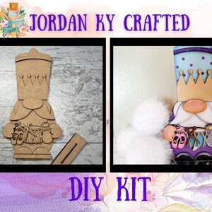 May include: A DIY craft kit featuring a wooden gnome with a crown, holding a pair of pink ballet shoes and a musical instrument. The gnome is painted in shades of blue, purple, and gold. The kit includes all the necessary materials to create the gnome, including the wooden pieces, paint, and brushes. The kit is labeled "JORDAN KY CRAFTED DIY KIT".