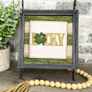 May include: A framed wooden sign with the word "LUCKY" in gold lettering and a green shamrock. The sign has a black frame and a green and white striped background. A wooden bead garland and a plant are in the foreground.