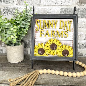 May include: A decorative sign with the words "SUNNY DAY FARMS" in yellow letters. The sign features three yellow sunflowers with brown centers. The sign is in a black frame and stands on a wooden surface.