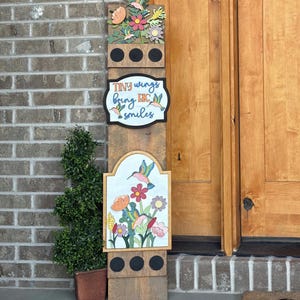 May include: A wooden sign with a hummingbird and flower design. The sign has the text "Tiny wings bring BIG smiles". The sign is made of wood and has a rustic look. The sign is decorated with colorful flowers and hummingbirds.