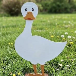 May include: A white duck-shaped garden ornament with a yellow-orange beak and feet. The duck has large black eyes with white highlights. It stands on a rectangular brown base featuring the text #smilecoproductions printed in white lettering.