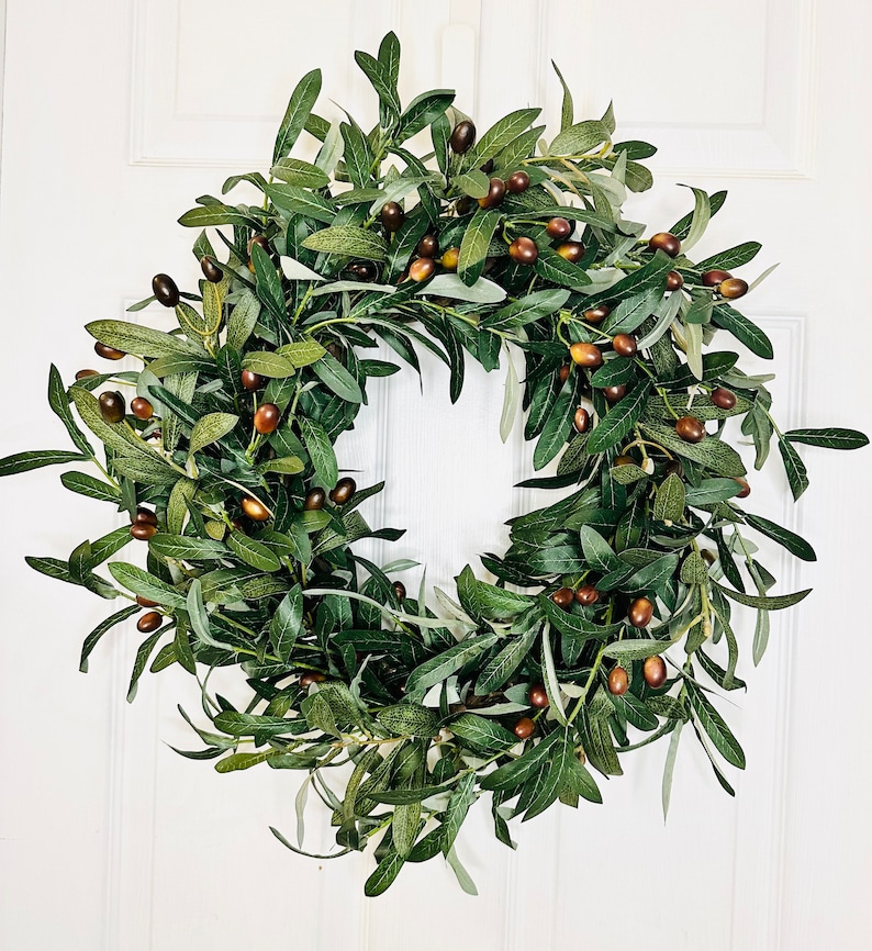 May include: A green olive branch wreath with brown olives. The wreath is made of artificial olive branches and is perfect for adding a touch of nature to your home decor.
