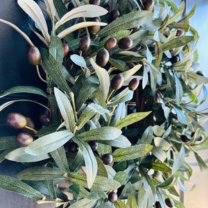 May include: A green olive branch wreath with dark green leaves and black olives. The wreath is hanging on a dark blue door.
