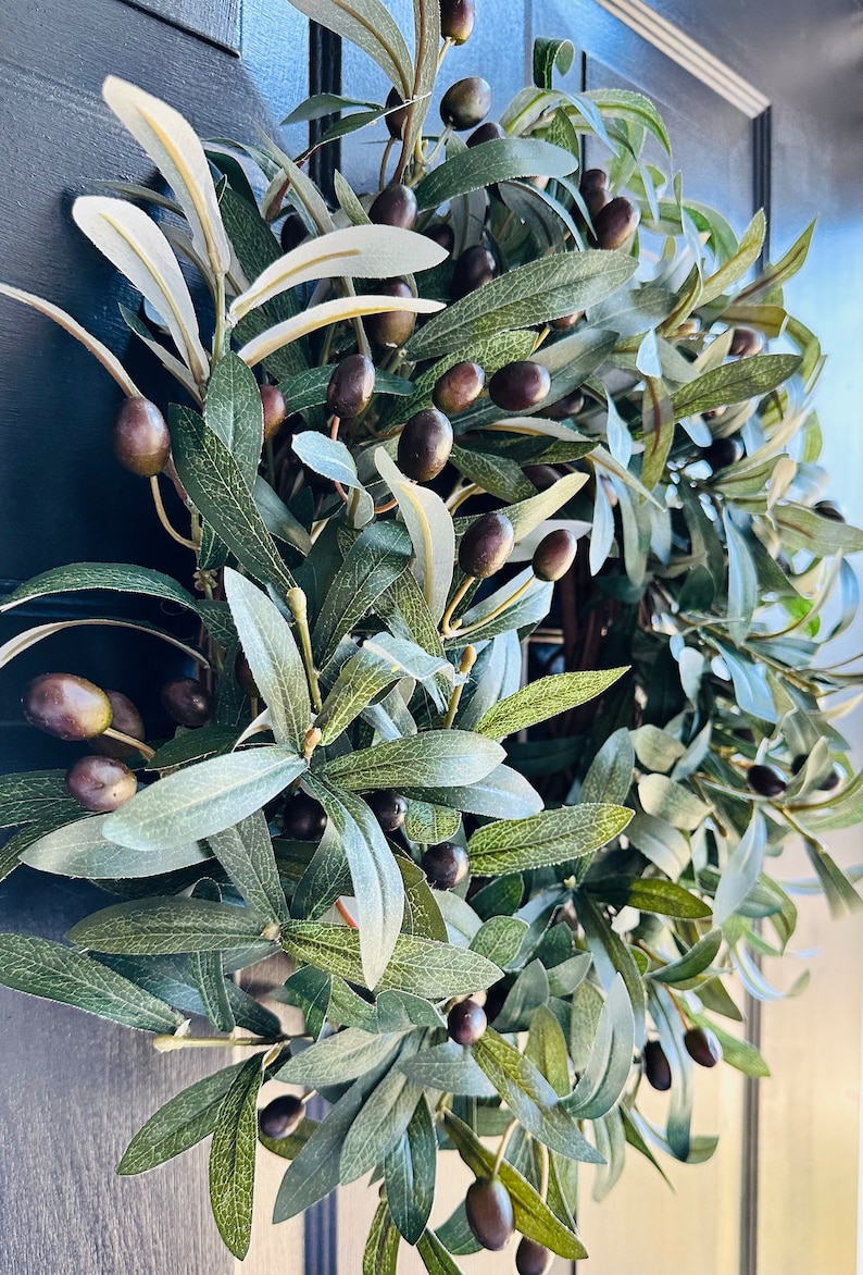 May include: A green olive branch wreath with dark green leaves and black olives. The wreath is hanging on a dark blue door.
