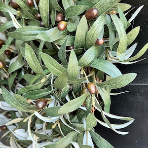 May include: A green olive branch wreath with brown olives. The wreath is made of artificial leaves and is perfect for adding a touch of nature to your home decor.