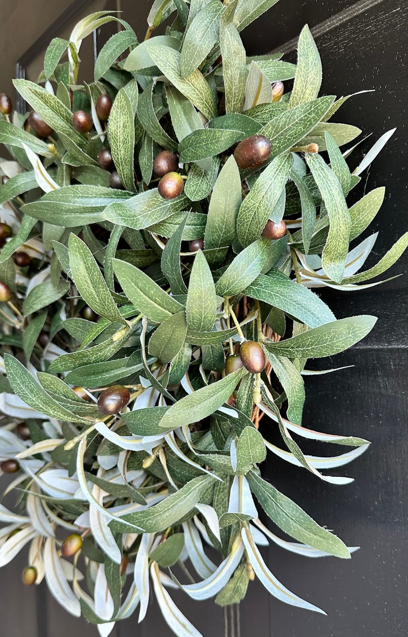 May include: A green olive branch wreath with brown olives. The wreath is made of artificial leaves and is perfect for adding a touch of nature to your home decor.