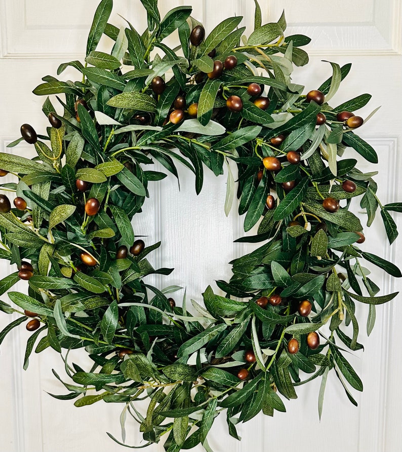 May include: A green olive branch wreath with brown olives. The wreath is made of artificial greenery and is perfect for decorating a door or wall.