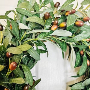 May include: A green olive branch wreath with brown olives. The wreath is made of artificial olive branches and is perfect for adding a touch of nature to your home decor.