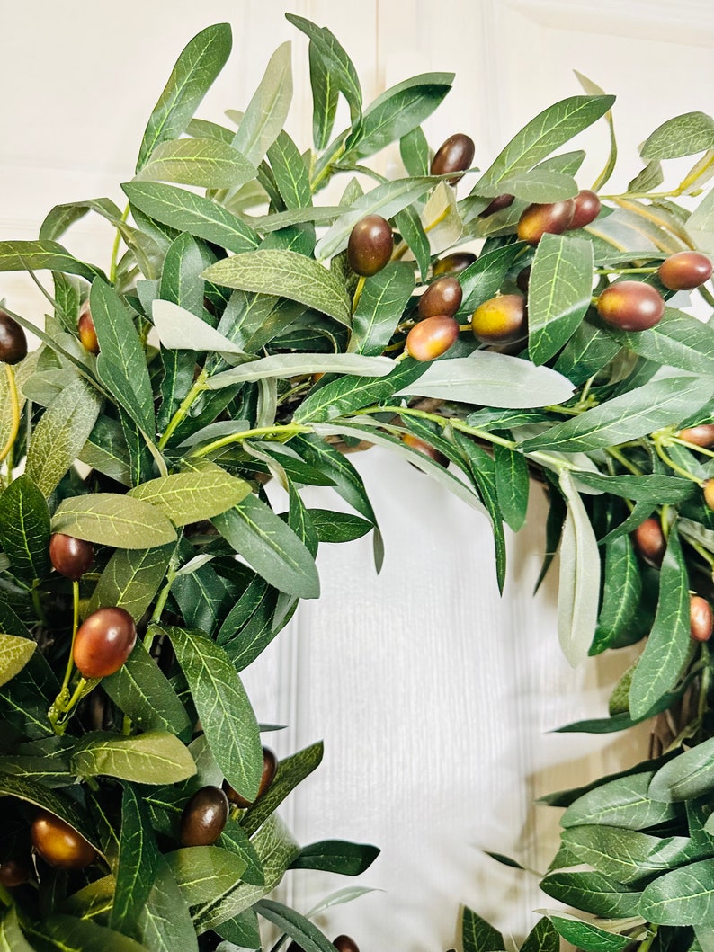 May include: A green olive branch wreath with brown olives. The wreath is made of artificial olive branches and is perfect for adding a touch of nature to your home decor.