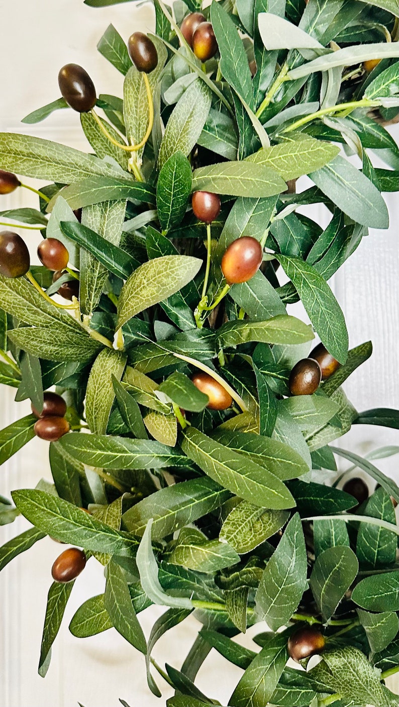 May include: A close-up of a green olive branch wreath with brown olives. The wreath is made of artificial leaves and olives.